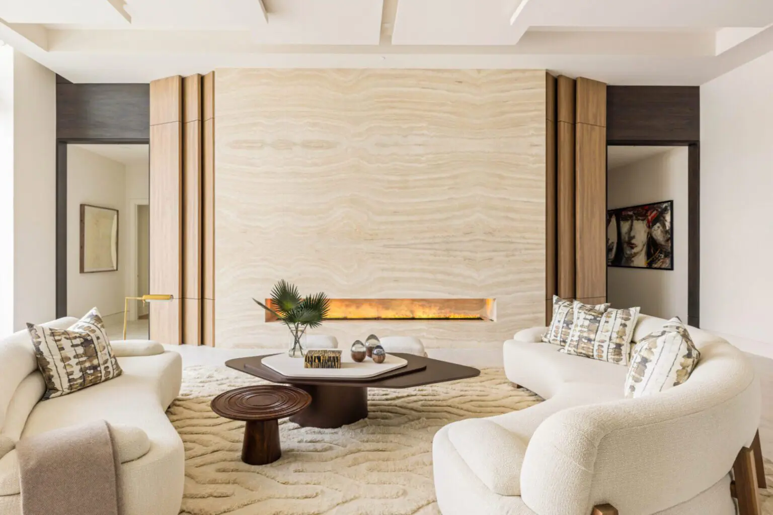 Modern living room with two cream sofas, a textured beige wall with a linear fireplace, abstract artwork, and a central coffee table with decorative objects.