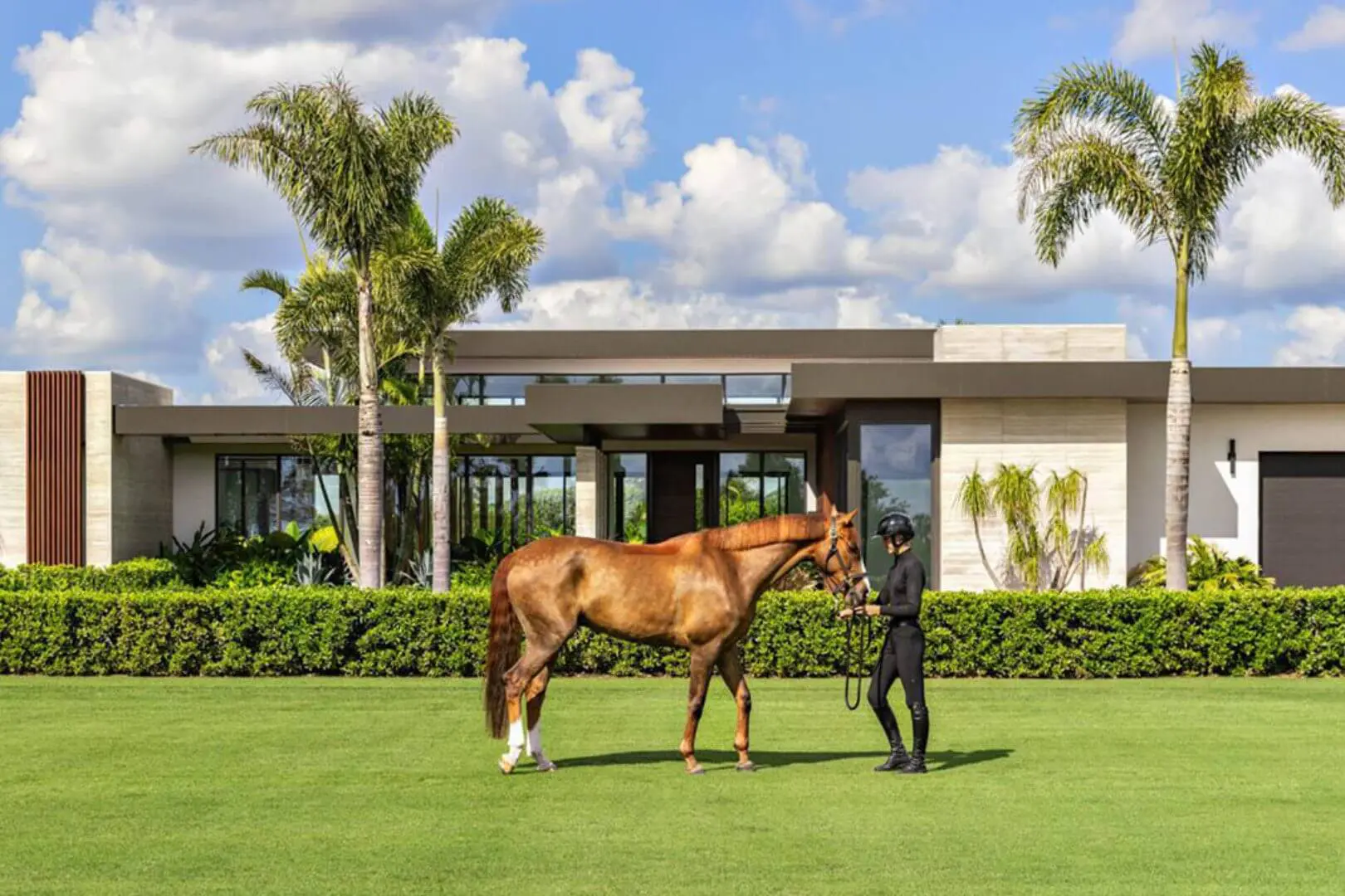 Woman standing next to a brown horse in front of a luxurious villa in Miami, surrounded by lush green grass