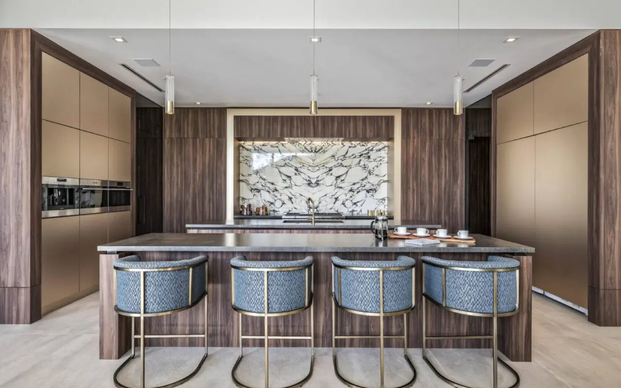 Modern kitchen with a marble backsplash, wood cabinetry, gold accents, an island with four blue upholstered barstools, and built-in appliances.