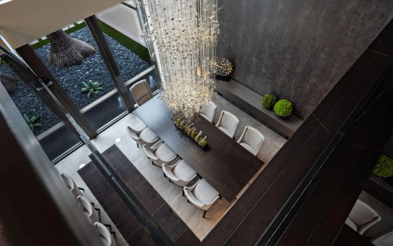 A modern dining room with a long dark table, twelve light-colored chairs, a tall chandelier, and large windows overlooking a minimalist garden.