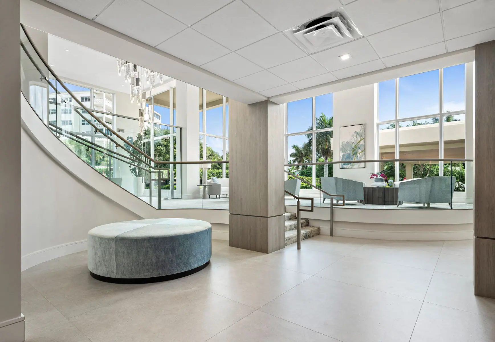 Modern lobby with a round ottoman, curved staircase, large windows, and a seating area with chairs and a table near tall indoor plants. Natural light fills the space.