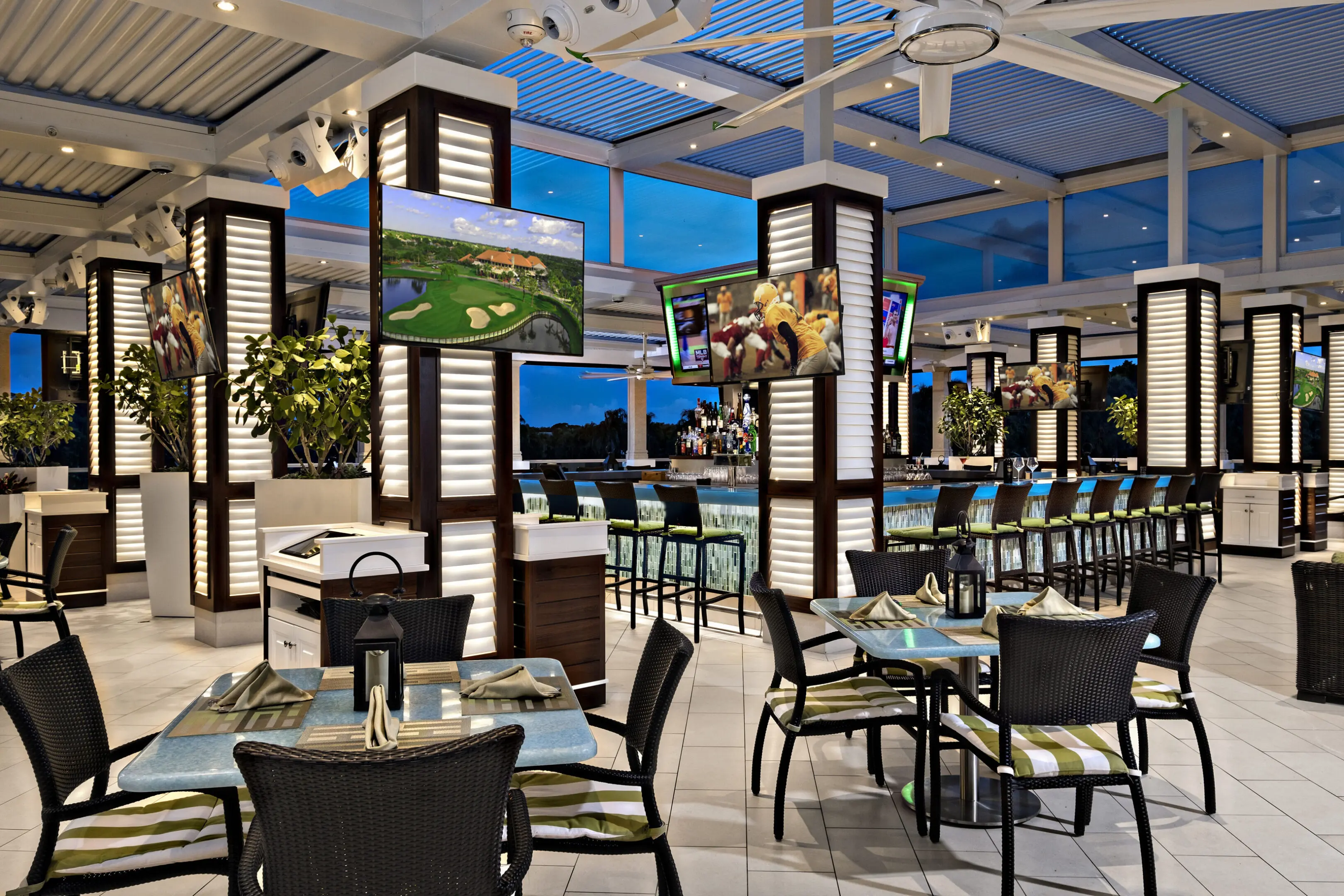 Modern restaurant interior with striped chairs, neatly set tables, large column light fixtures, multiple ceiling fans, and several flat-screen TVs displaying sports and golf.