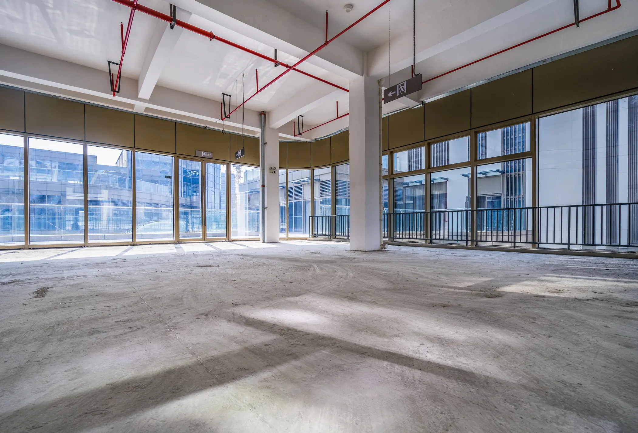 Empty commercial space with large floor-to-ceiling windows, concrete floor, exposed ceiling pipes, and ample natural light.