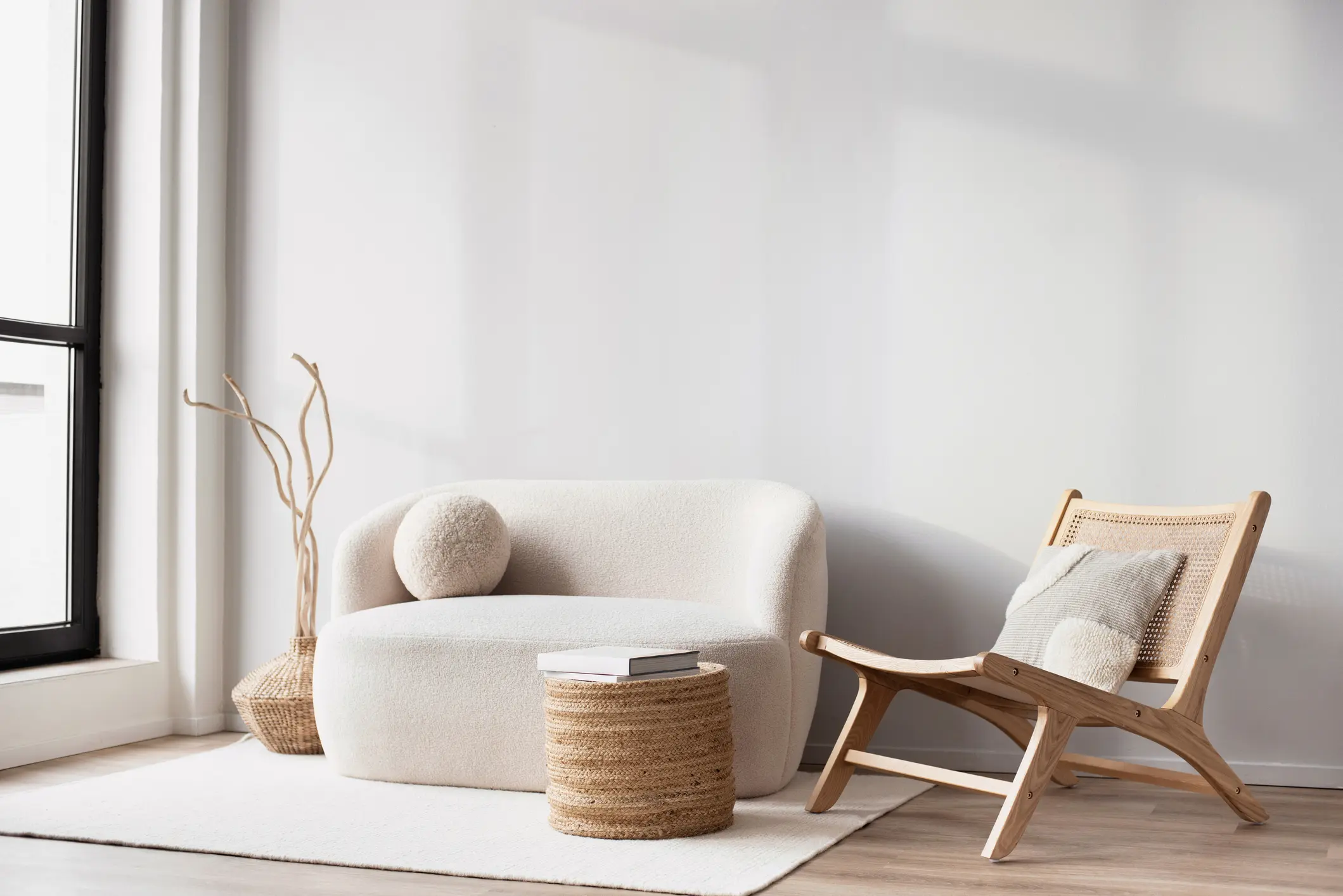 Minimalist living room with a white sofa, wooden lounge chair, wicker side table, decorative branch, and soft natural light.
