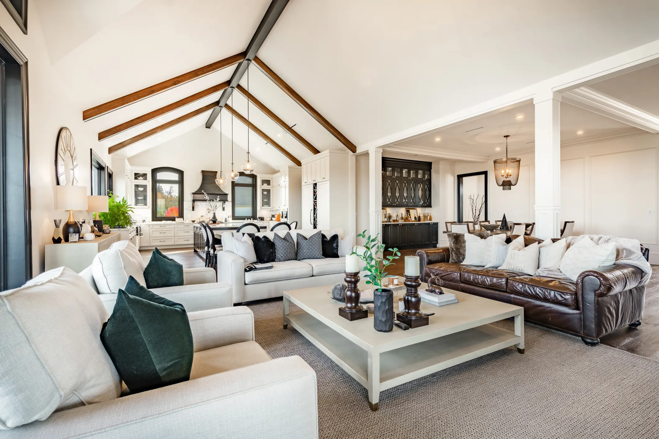 Spacious living room with vaulted ceiling, exposed beams, neutral sofas, a leather couch, coffee table, and open view to kitchen and dining area with modern decor.