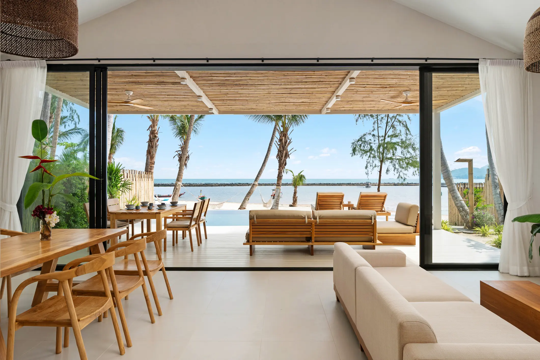 Open living and dining area with wooden furniture, leading to a deck with loungers overlooking a body of water and palm trees under a clear sky.