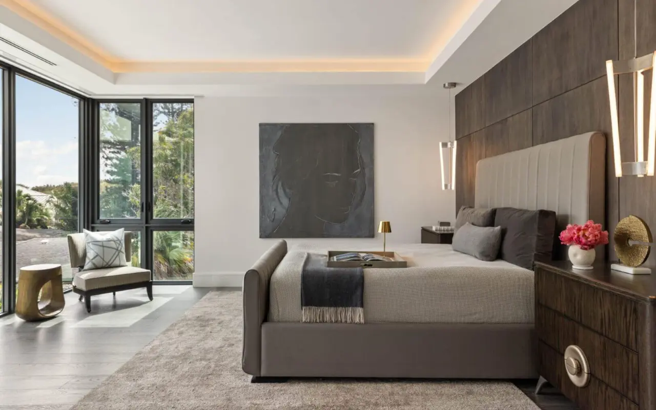 Modern bedroom with a large bed, dark wood paneling, two nightstands, a chair by tall windows, and artwork on the wall. Neutral and grey tones dominate the decor.