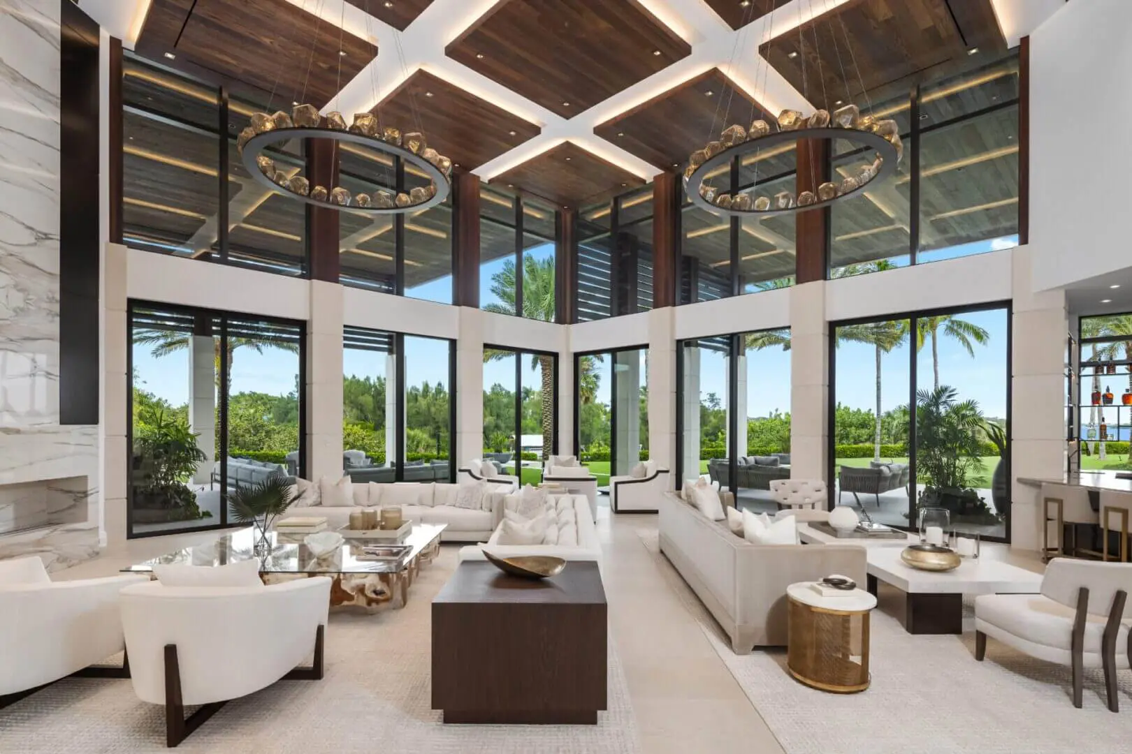 Spacious modern living room with floor-to-ceiling windows, marble accents, neutral-colored furniture, large circular chandeliers, and views of greenery outside.