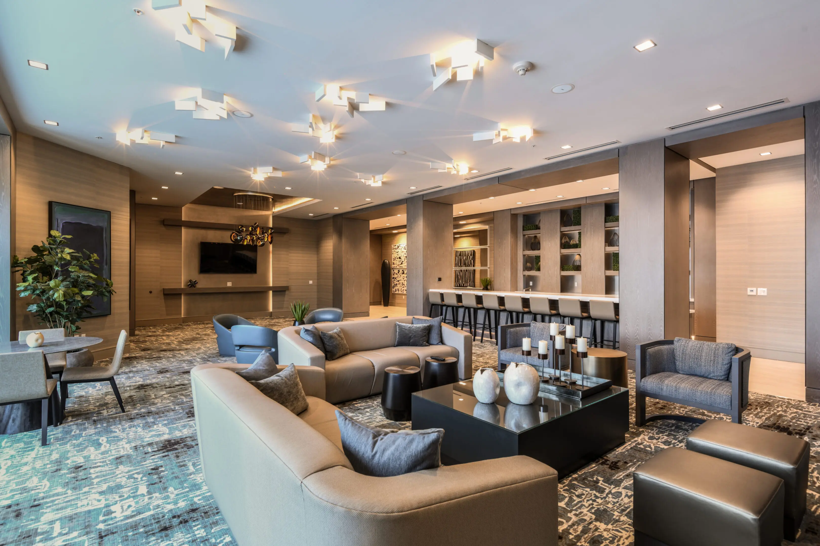 Modern lounge area with sofas, armchairs, coffee tables, decorative lighting, wall art, large TV, plants, and a bar with stools. Neutral colors and contemporary decor create a spacious, stylish setting.