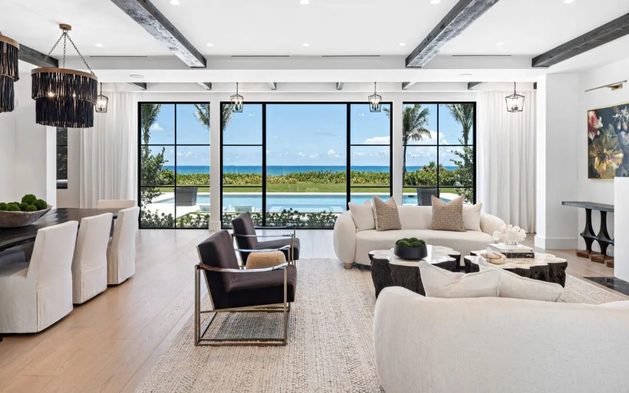 Modern living and dining area with neutral furniture, large windows, ocean view, and pool outside; black accents and ceiling beams add contrast to the bright, open space.