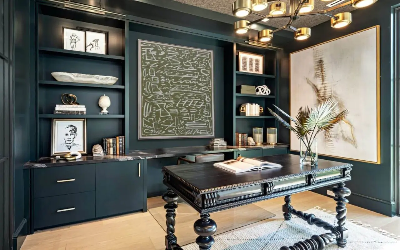 Modern study with dark built-in shelves, abstract wall art, decorative objects, books, and a large ornate desk with an open book. A pendant light hangs from the ceiling.