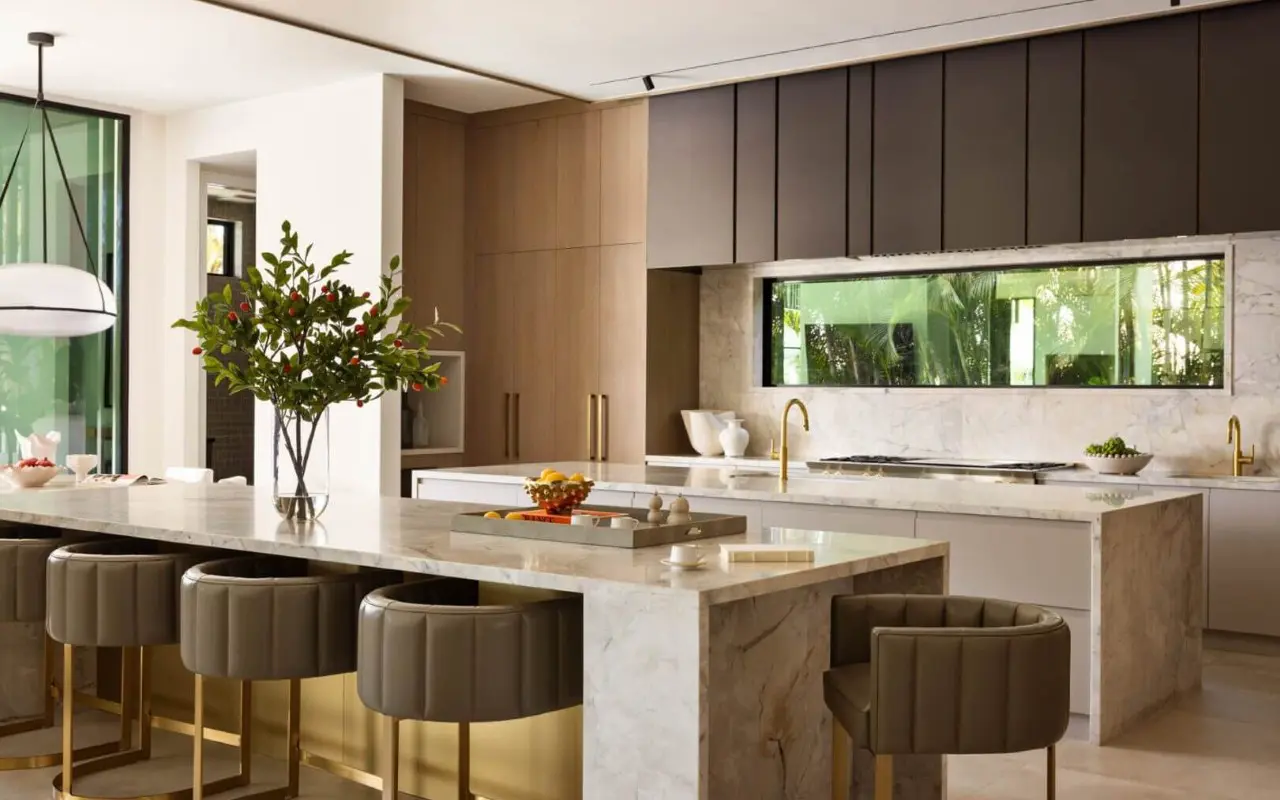 Modern kitchen with marble countertops, high-back stools, wood and dark cabinets, large window, and decorative vase with greenery on the island.