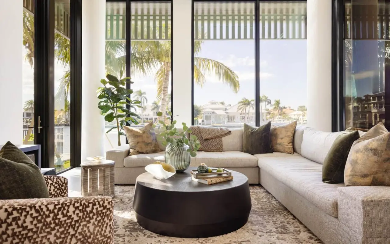 Modern living room with a beige sectional sofa, patterned armchair, round black coffee table, and large windows showing palm trees and houses outside.