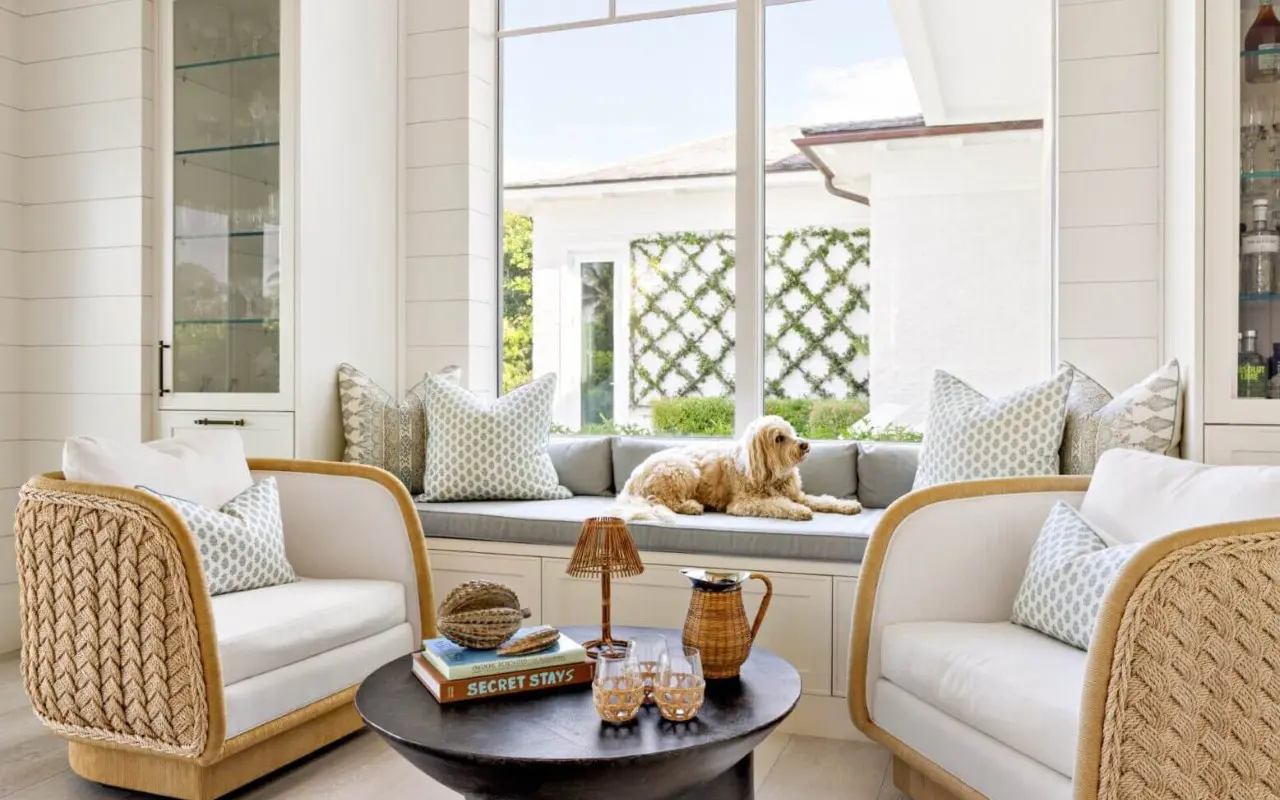 A dog lies on a window bench with cushions in a bright, modern living room featuring two armchairs, a round coffee table, and large windows.