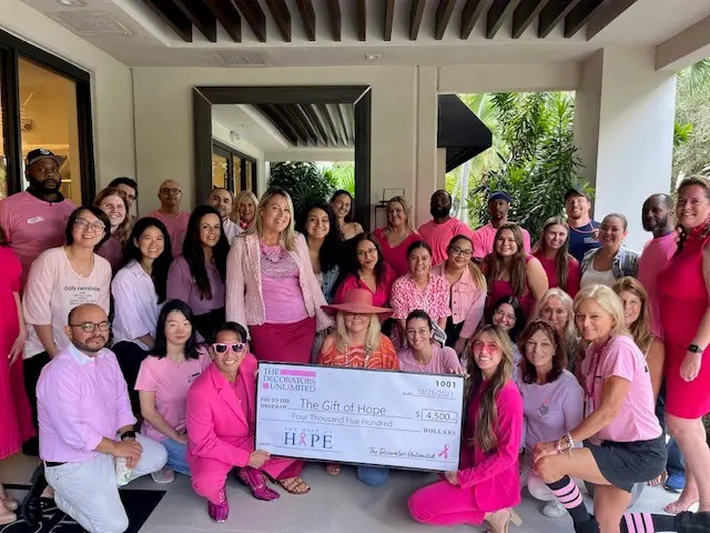A group of people dressed in pink pose together indoors, holding a large check for $4,500 made out to “The Gift of Hope.”.
