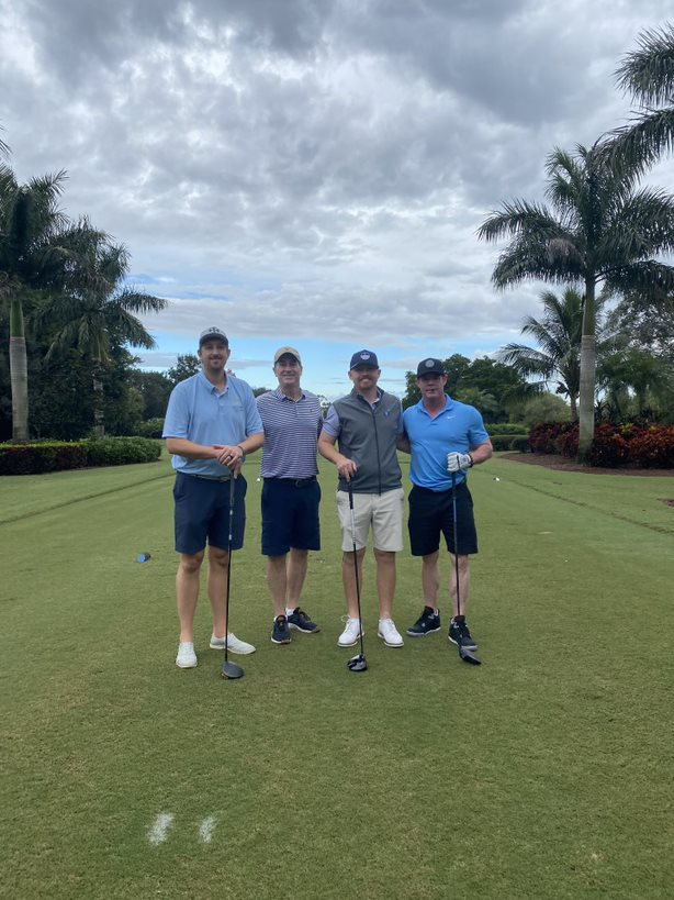 Four men holding golf clubs stand on a golf course tee box, surrounded by palm trees under a cloudy sky.