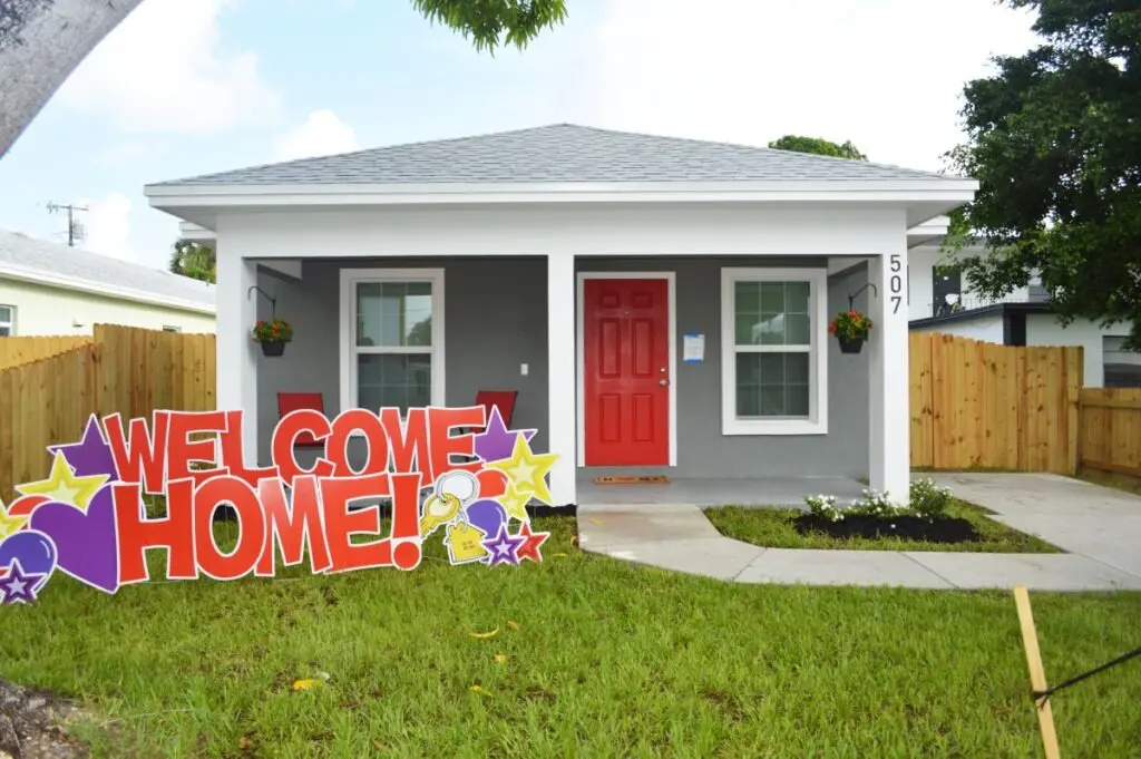 Gray house with red door, front porch, potted plants, and a large “Welcome Home!” sign on the lawn. House number 507 is visible on the porch post.
