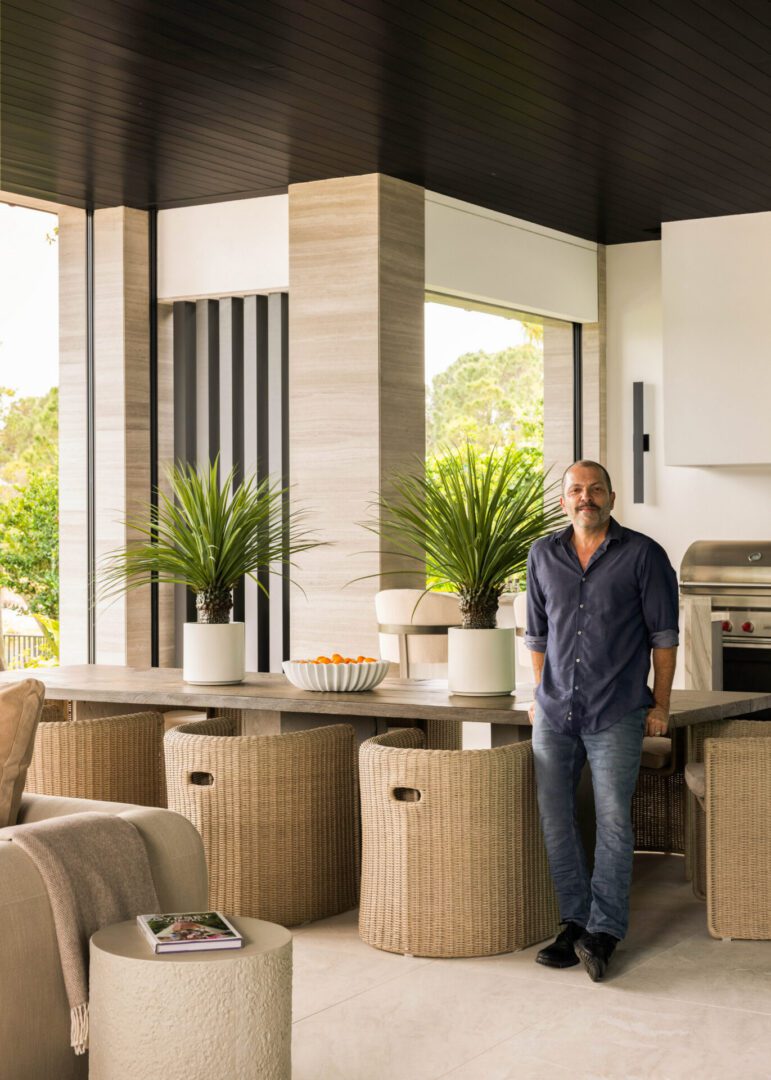 Man standing in front of a kitchen with a table, chairs, sofa, pillows, cloth, book, plants, bowl, and other objects