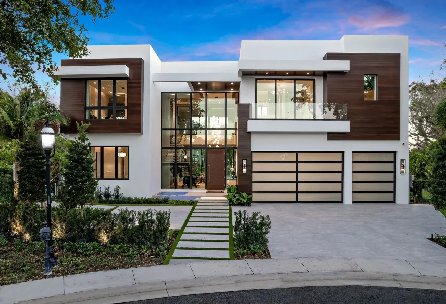 Modern two-story house with large glass windows, wood and white exterior, double garage, front garden, and paved driveway at sunset.