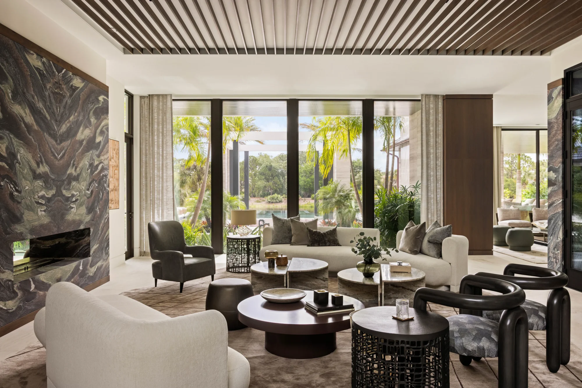 Modern living room with neutral-toned furniture, round coffee tables, large windows, and views of a tropical garden outside.
