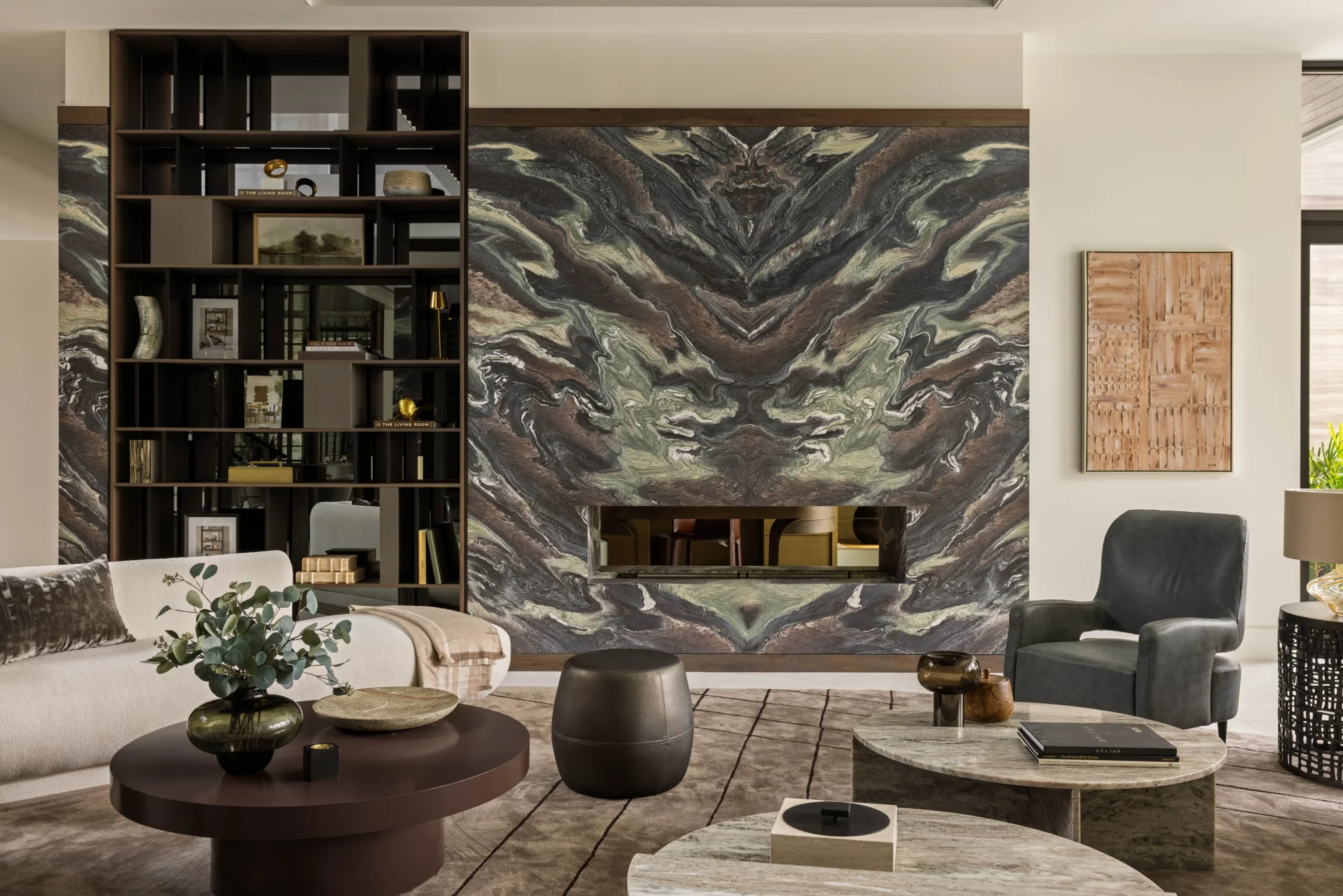 Modern living room with abstract stone fireplace, open shelves with books and decor, neutral furniture, and round tables on a patterned rug.