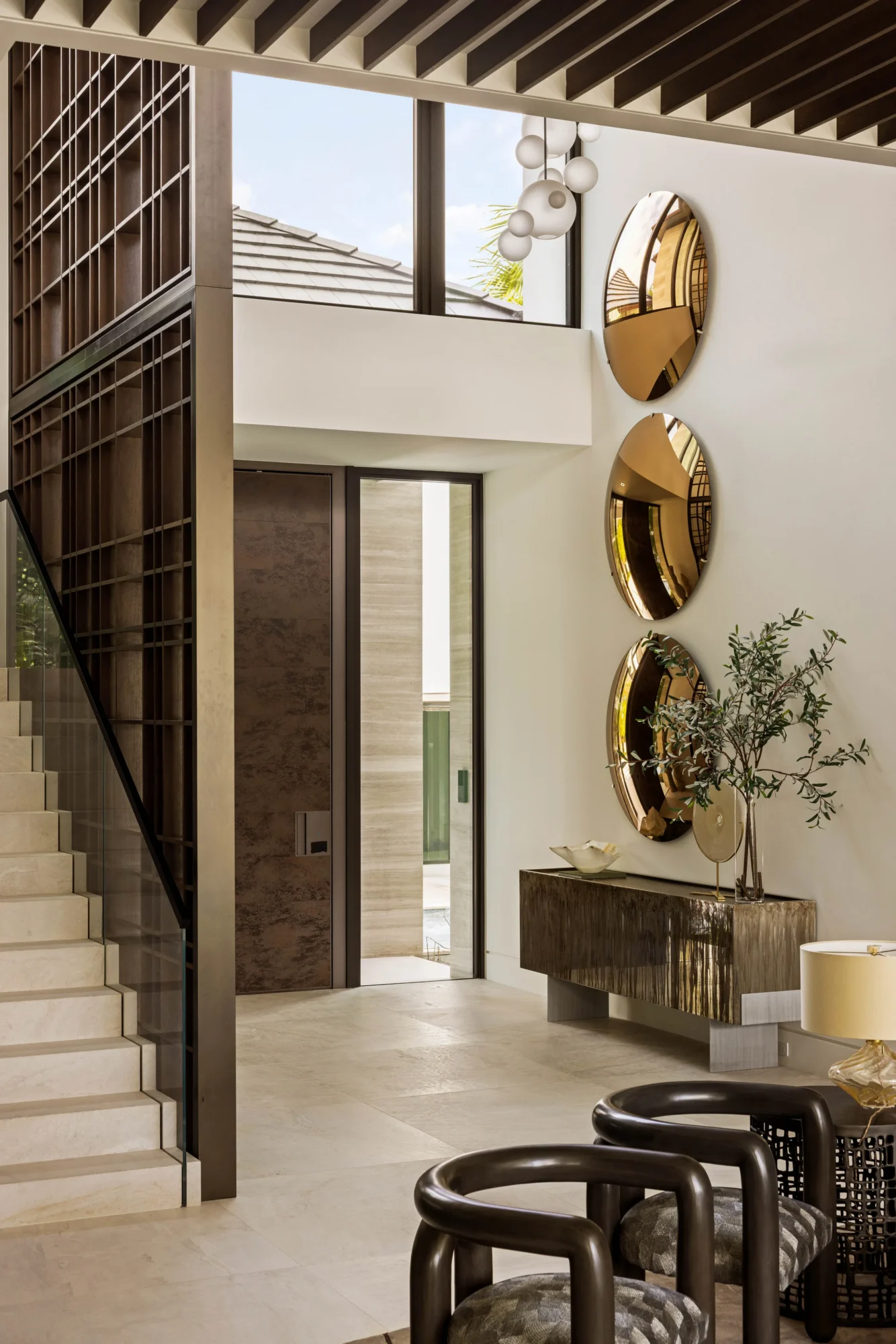 Modern entryway with a staircase, large windows, three round gold wall mirrors, a console table with a plant, and contemporary chairs.