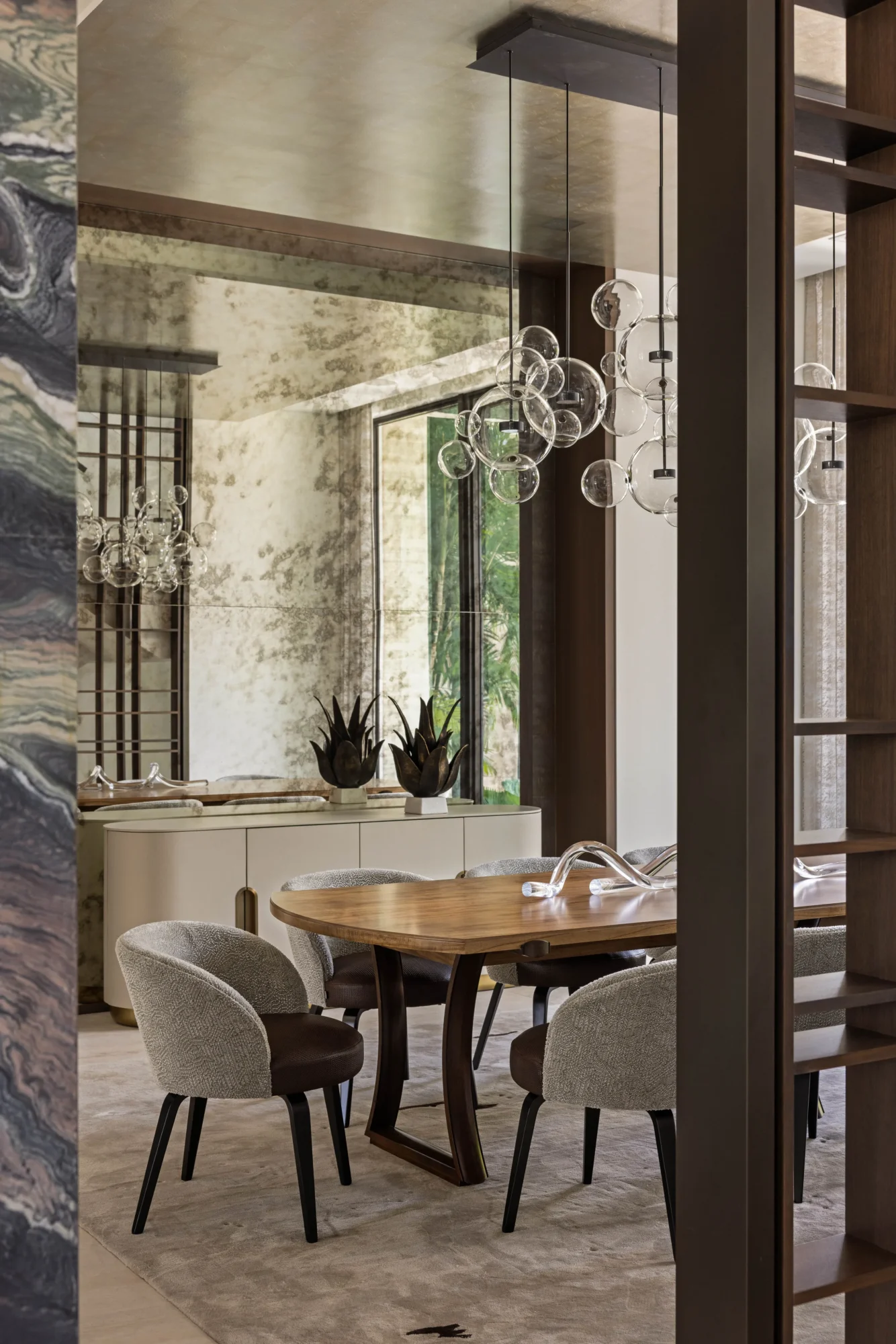 Modern dining room with a wooden table, four upholstered chairs, abstract glass chandelier, large mirror, and decorative plants near a window. Neutral tones and contemporary design elements are present.