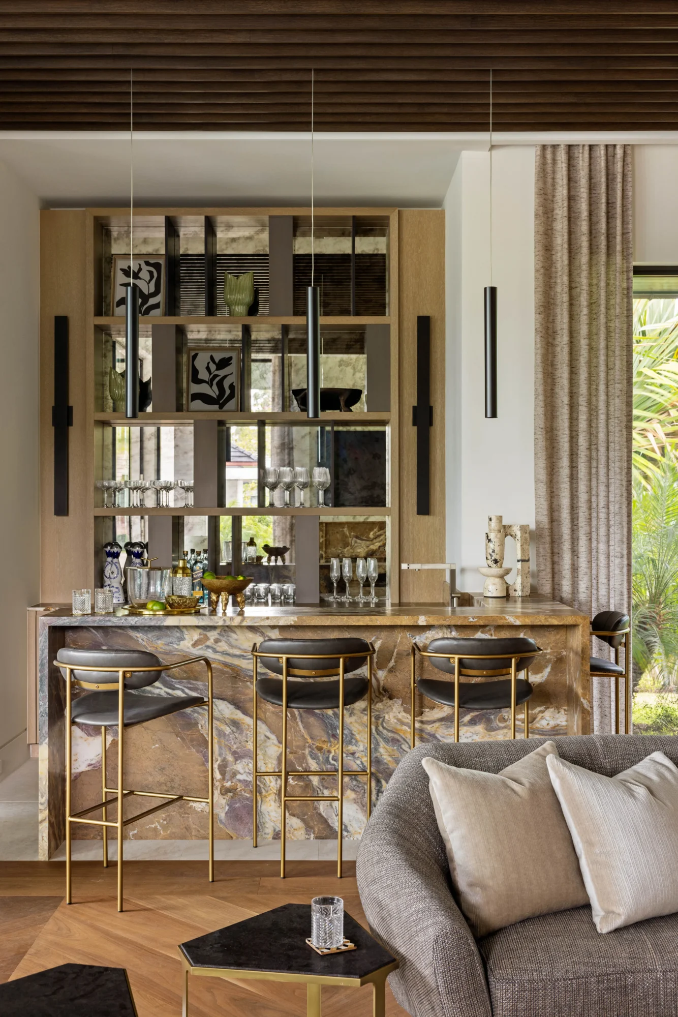 A modern home bar with a marble counter, gold barstools, glass shelves with glassware, and decorative items; a gray sofa and a window with curtains are in the foreground.