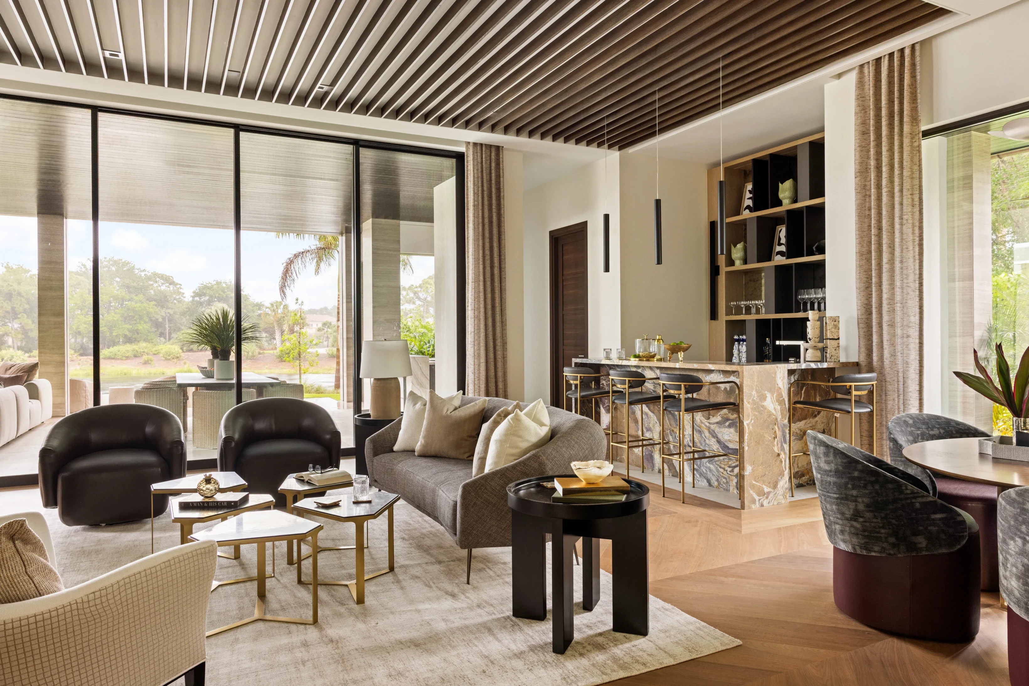 Modern open-concept living room and kitchen with large windows, neutral-toned furniture, marble surfaces, and wood accents, overlooking a green outdoor view.