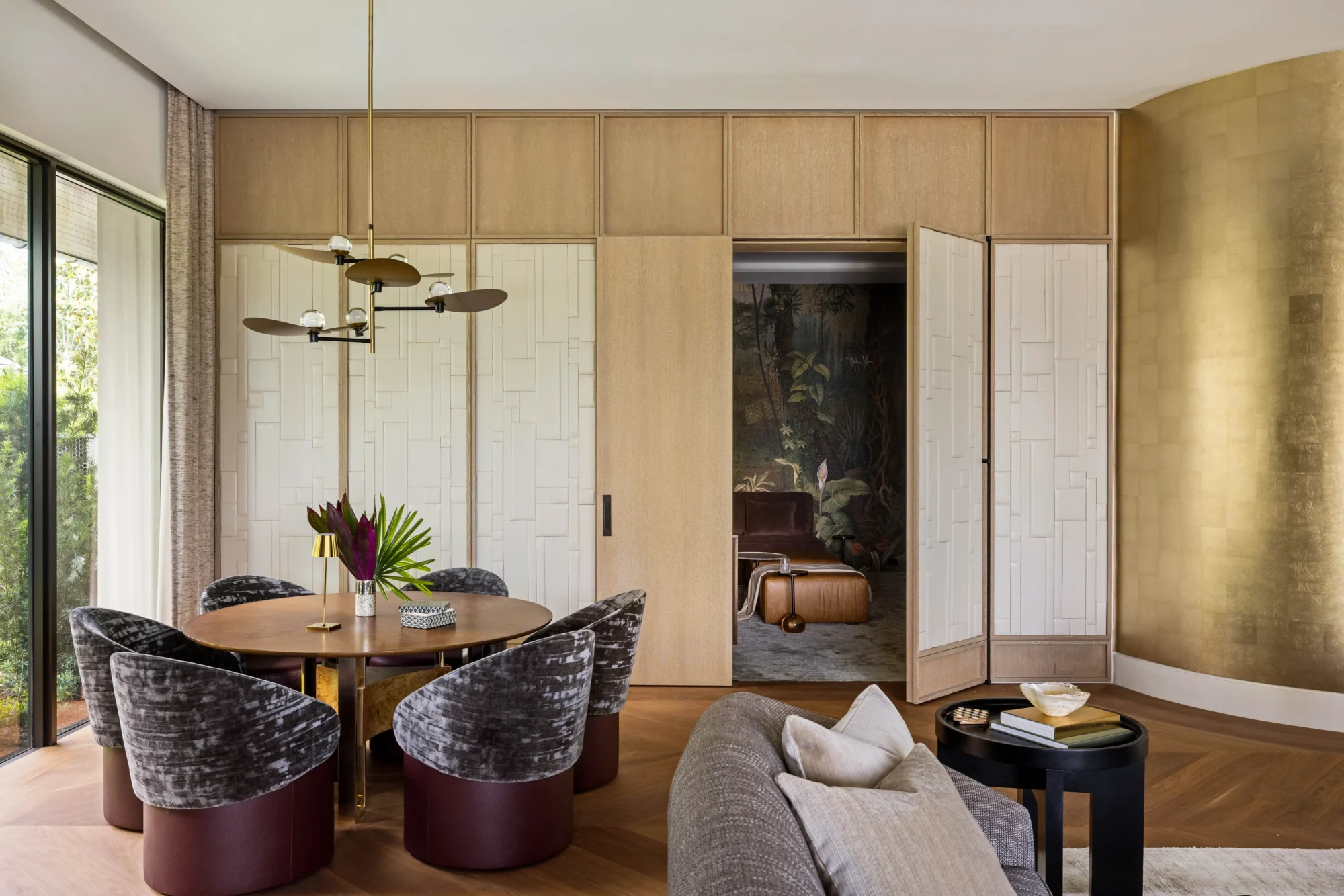 Modern dining area with a round table, four velvet chairs, a beige sofa, gold pendant light, wooden paneled wall, and double doors partially open to a room with a mural.