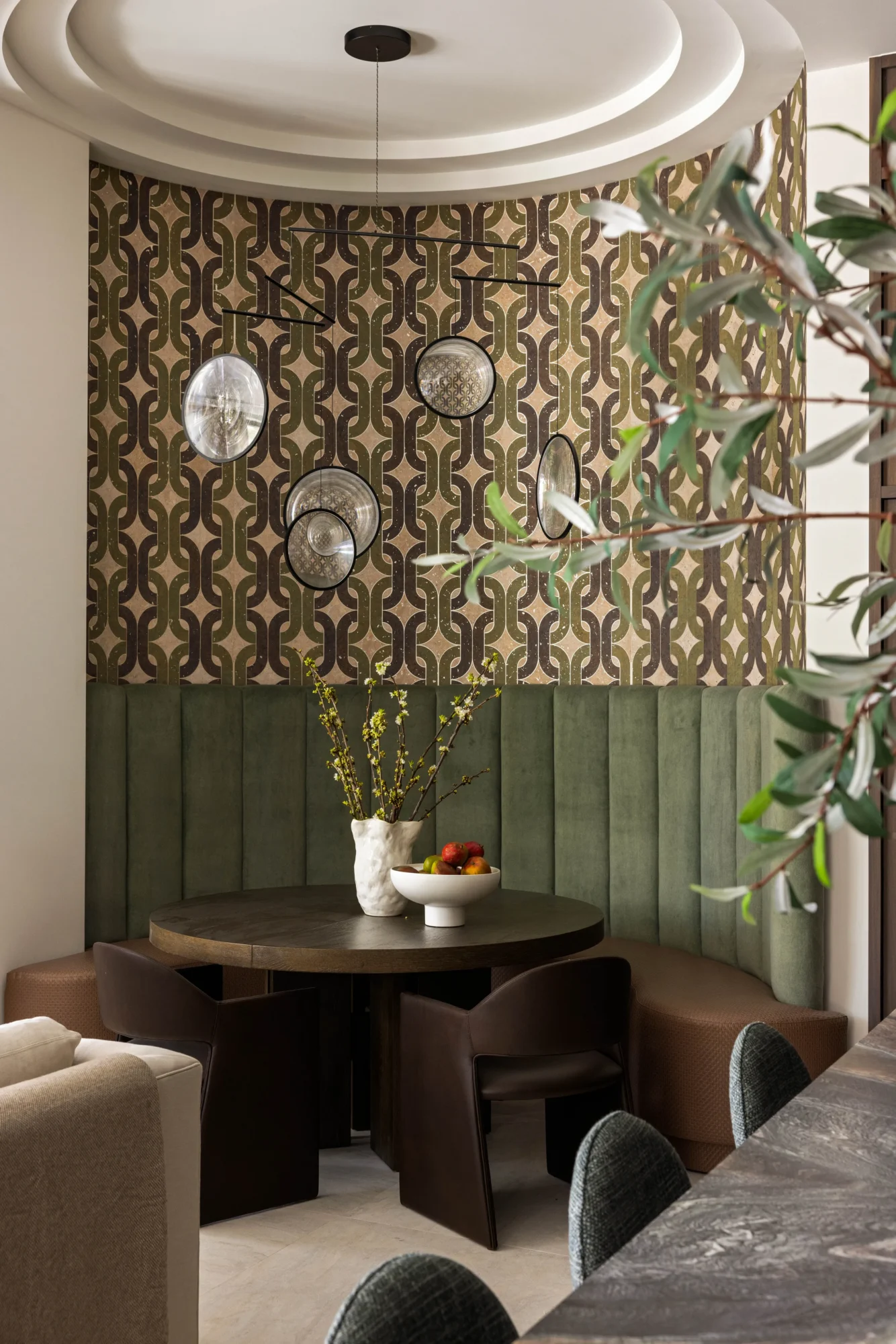 A modern dining nook with a round table, leather chairs, patterned wallpaper, and pendant lights; a vase with flowers and a fruit bowl sit on the table.
