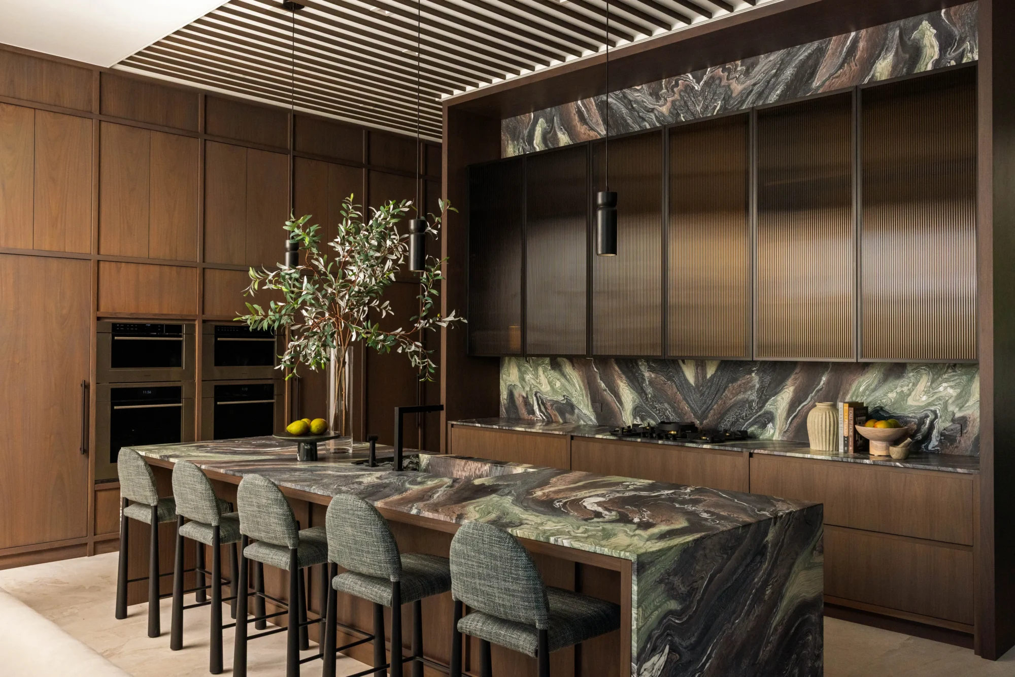 Modern kitchen with dark wood cabinets, marble-patterned island and backsplash, green cushioned chairs, pendant lights, and a vase with branches on the island.