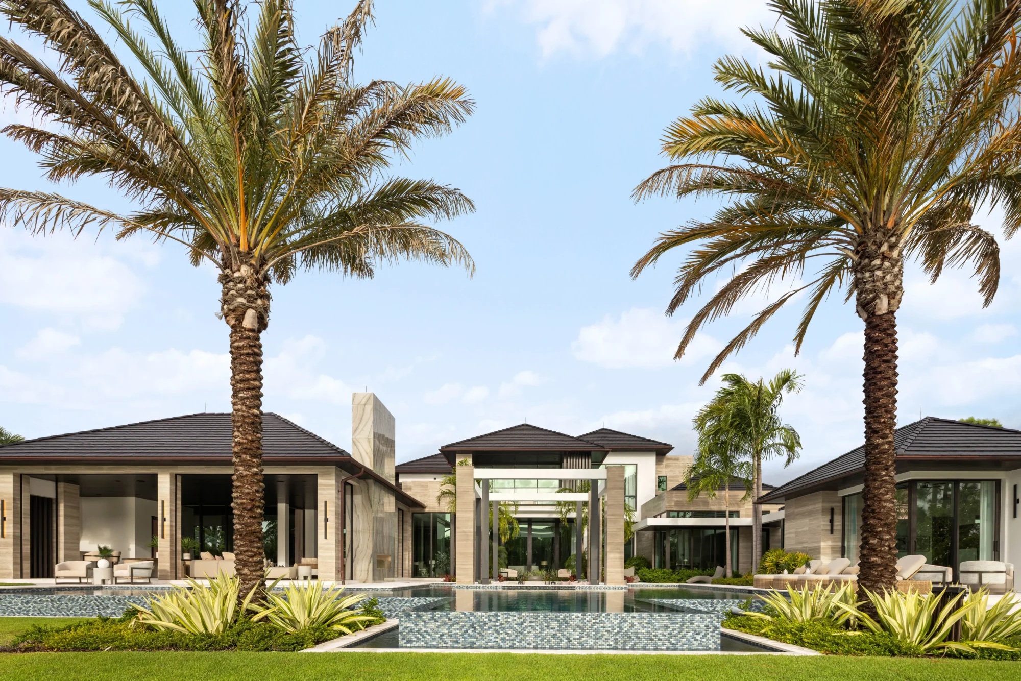 Modern luxury house with large glass windows, a central pool, and palm trees in the landscaped front yard under a blue sky.