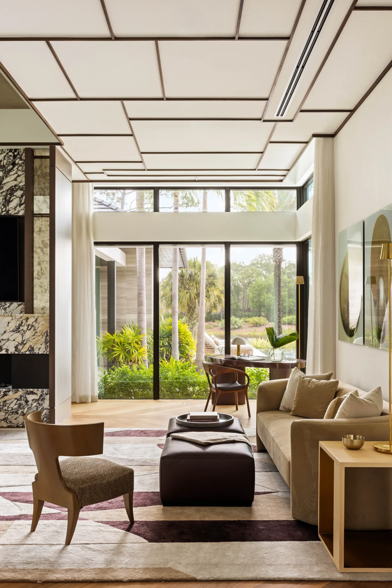 Modern living room with large windows, beige sofa, chairs, a geometric rug, ottoman, and a view of greenery outside. The space features a unique ceiling design and neutral color palette.