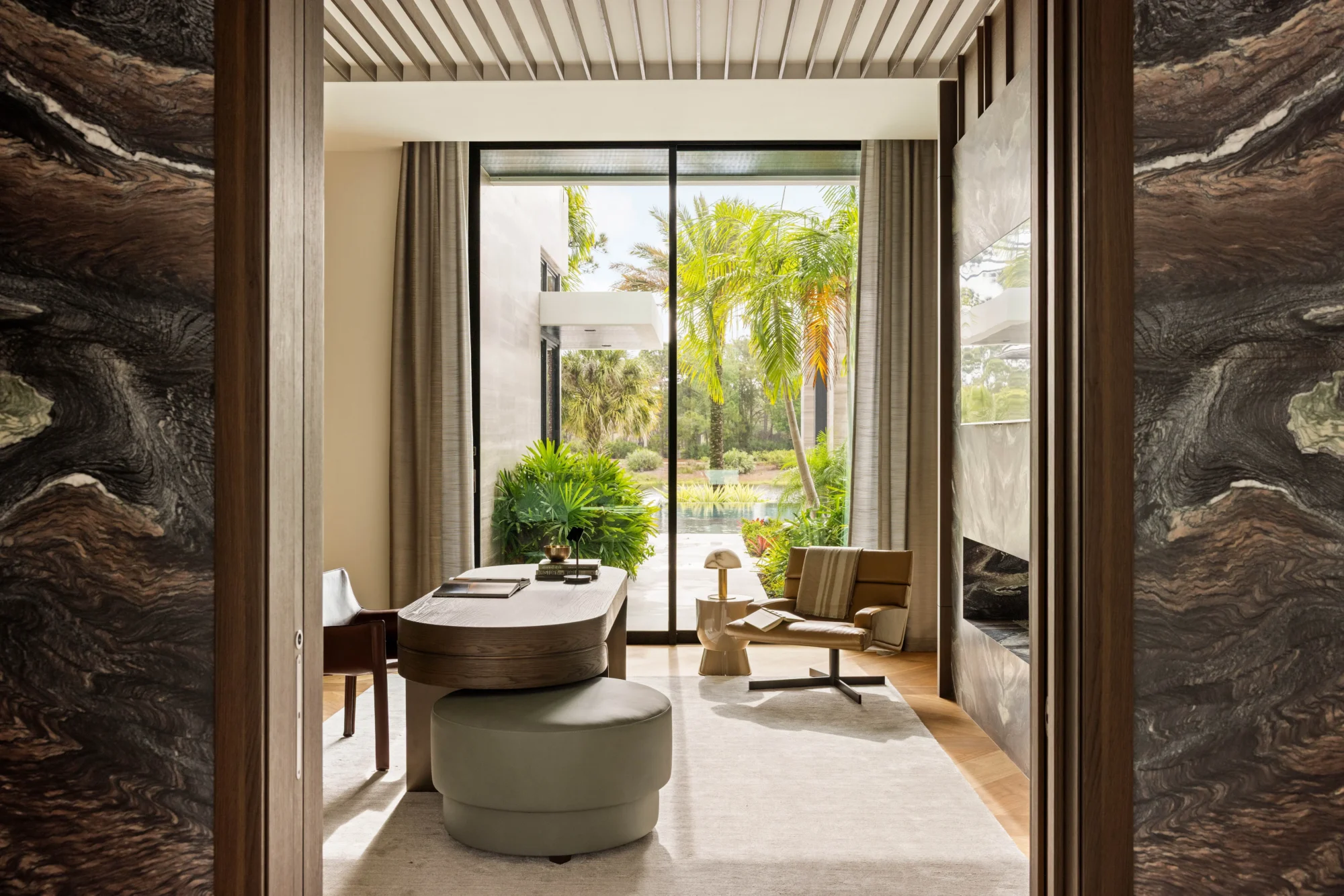 Modern home office with a curved desk, chair, and lounge chair near floor-to-ceiling windows overlooking a lush garden with palm trees.