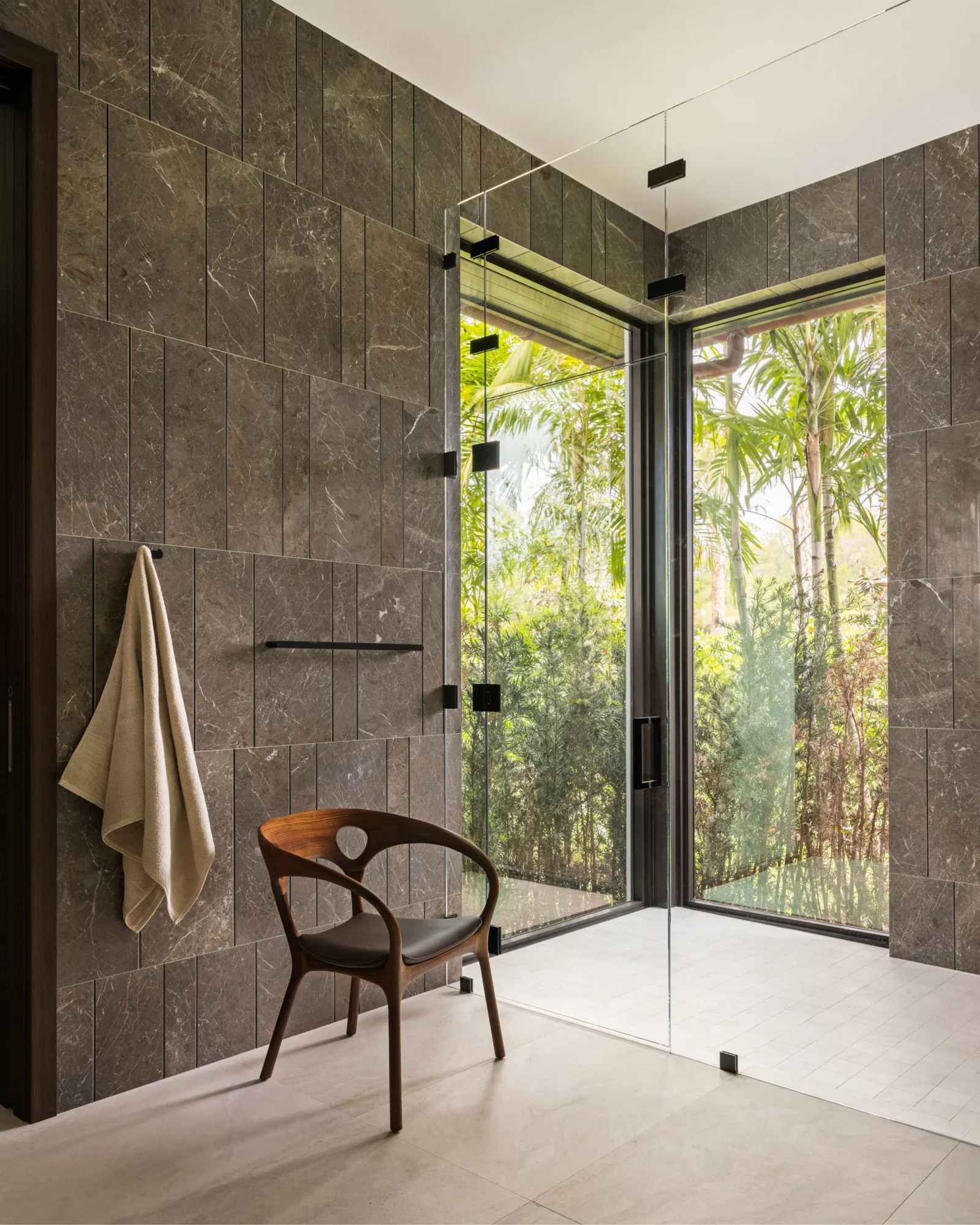 Modern bathroom with large glass shower, brown tiled walls, a wooden chair, a towel hanging on a hook, and large windows showing green plants outside.