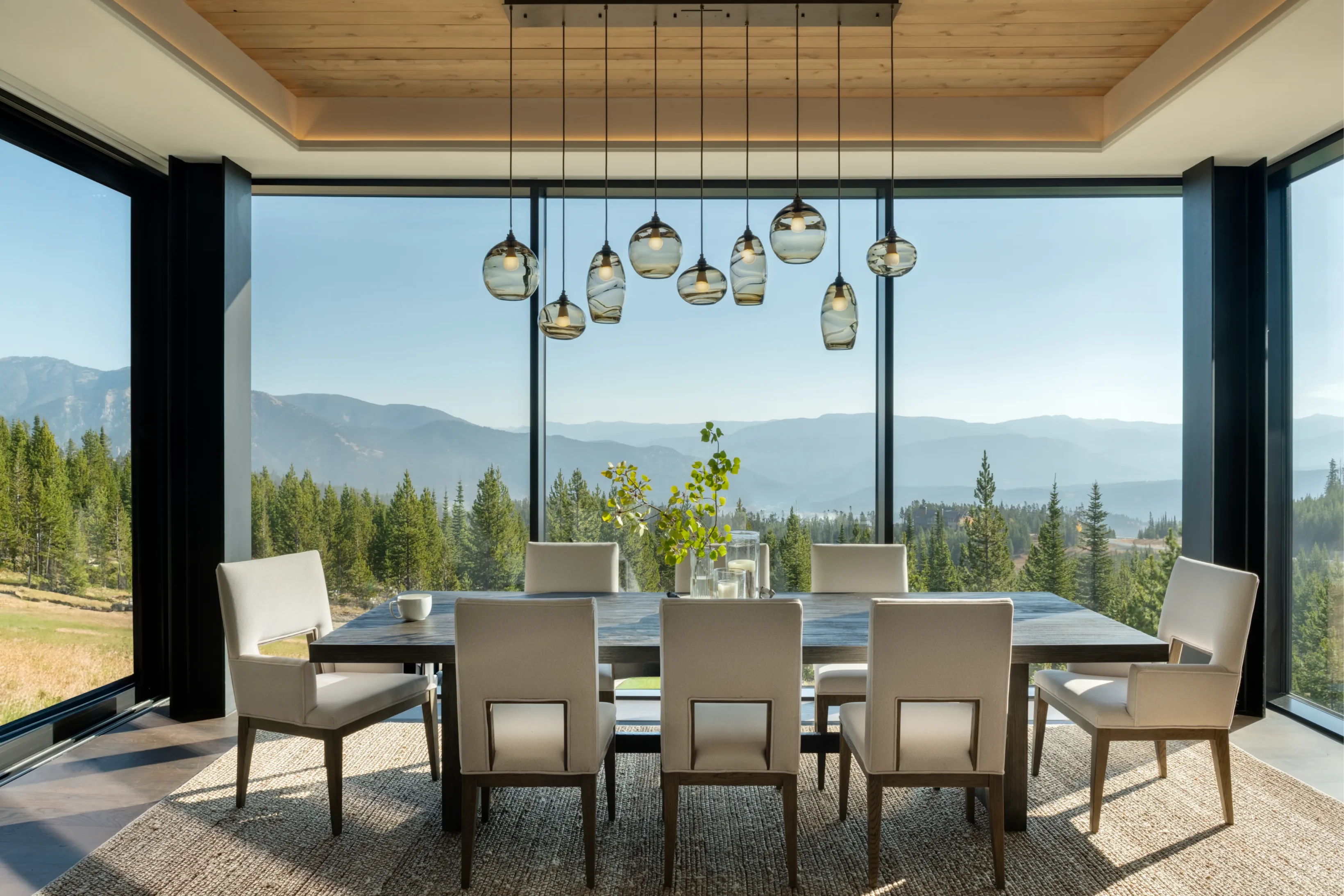 Modern dining room with large windows showing a mountain view, a rectangular table with eight chairs, pendant lights above, and a centerpiece with green branches.