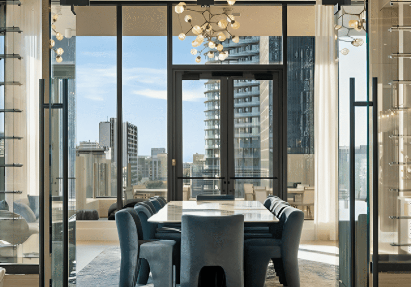 Modern dining room with blue chairs, large windows, city views, and a contemporary chandelier above a rectangular table. Glass doors and natural light enhance the spacious interior.